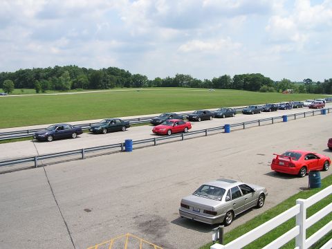 1993 Ford Taurus - A line of cars, including a silver 1993 Ford Taurus, are parked on a racetrack with a grassy field in the background.
