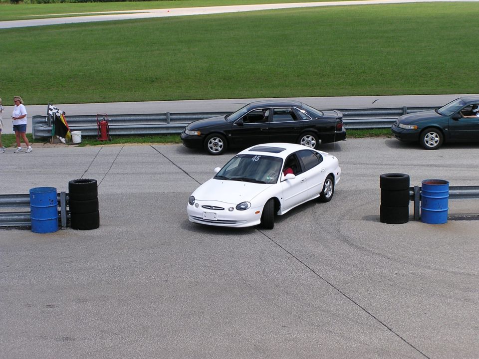 White Ford Taurus on a track, number 45 on windshield. Black car behind it, green car to the right
