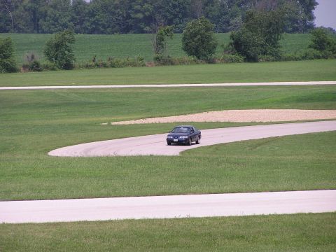 1993 Ford Taurus - A dark sedan navigates a curve on a race track surrounded by green fields and trees.