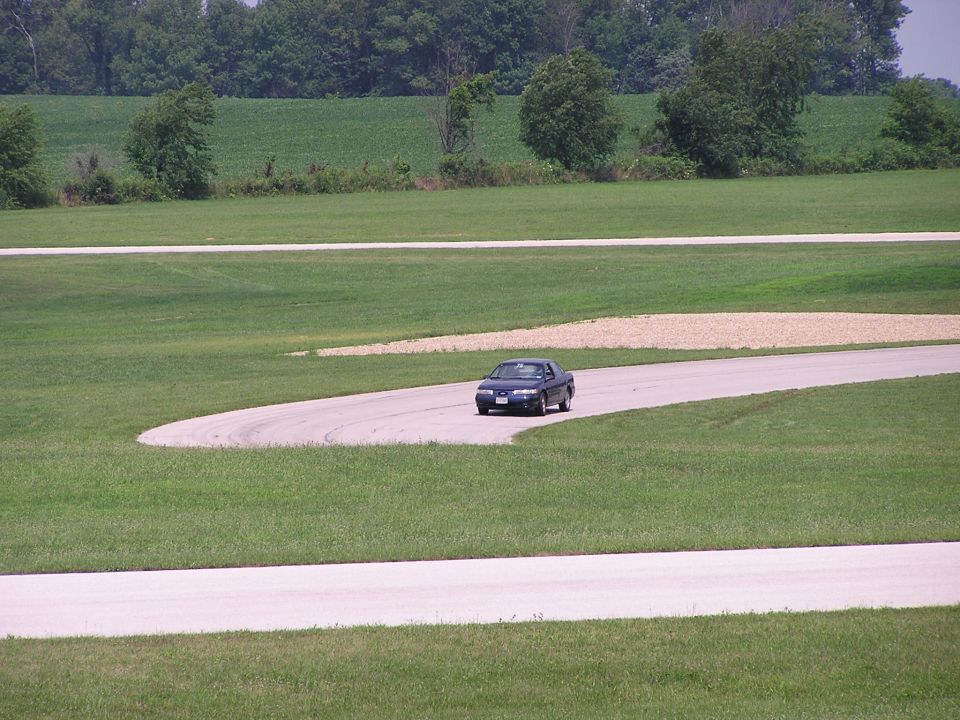dark sedan driving on a paved track with grassy fields and trees in the background