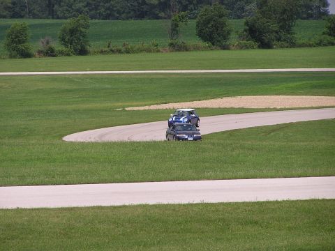 1993 Ford Taurus - Two cars navigate a turn on a paved racetrack surrounded by green grass and fields.