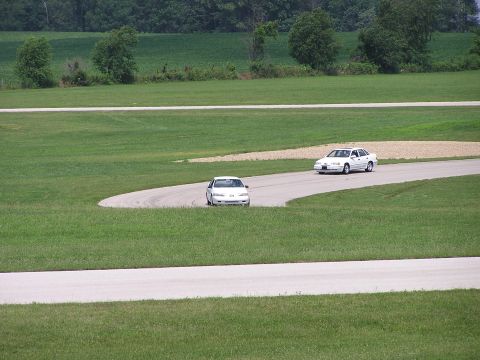 1993 Ford Taurus - Two white sedans navigate a turn on a grassy road course during an event.