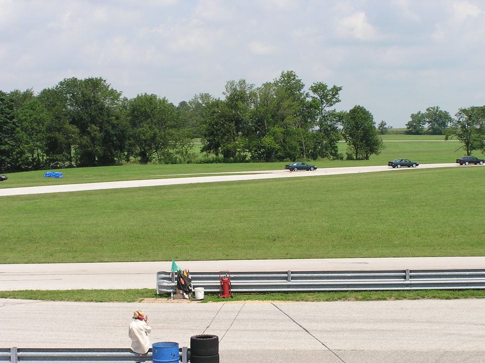 three dark sedans drive in a line on a paved track with grassy fields and trees in the background