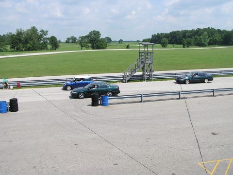 1993 Ford Taurus - Multiple vehicles on a race track with a control tower and grassy surroundings under a cloudy sky.