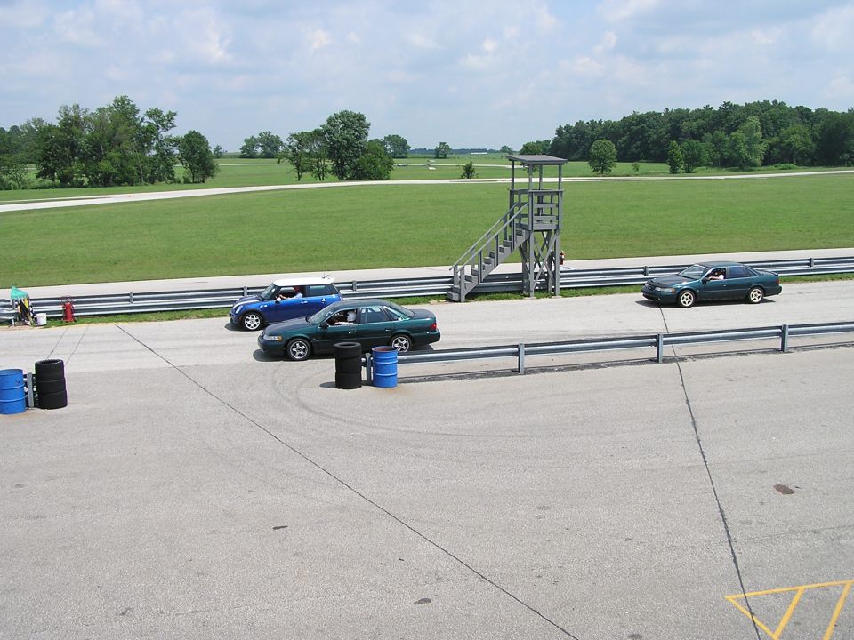 Race track with a green sedan and a blue hatchback, plus a distant car. A tower stands beside the track