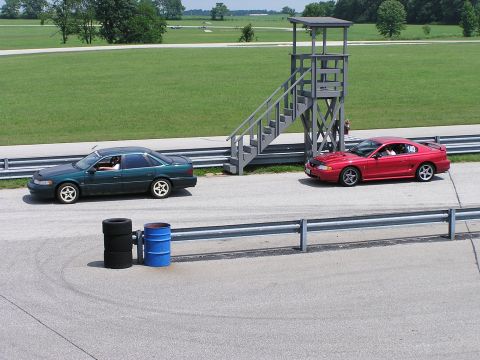 1993 Ford Taurus - Two cars, a green sedan and a red coupe, are on a paved track with a race control tower in the background.