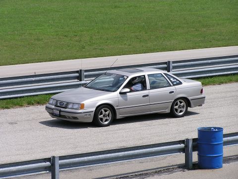 1993 Ford Taurus - A silver sedan drives on a paved track with a grassy area and guardrails visible.