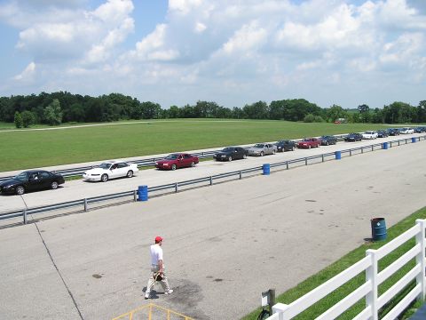 1993 Ford Taurus - A line of sedans is parked on the side of a racetrack under a partly cloudy sky.