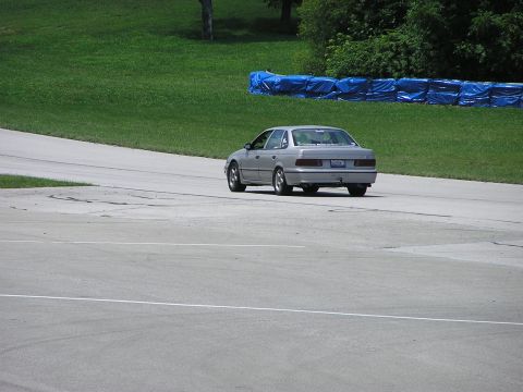 1993 Ford Taurus - A silver sedan navigates a turn on a paved track with grassy banks and blue safety barriers.