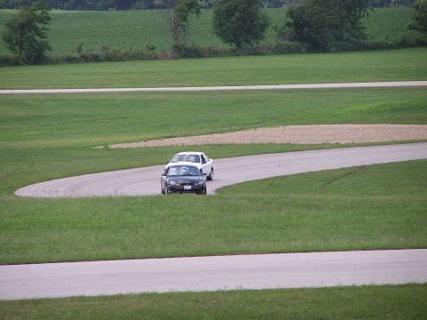 1993 Ford Taurus - Two sedans navigate a turn on a paved track surrounded by grassy fields.