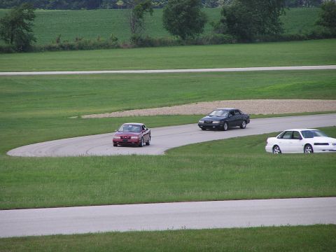 1993 Ford Taurus - Three sedans navigate a turn on a grassy race track under an overcast sky.