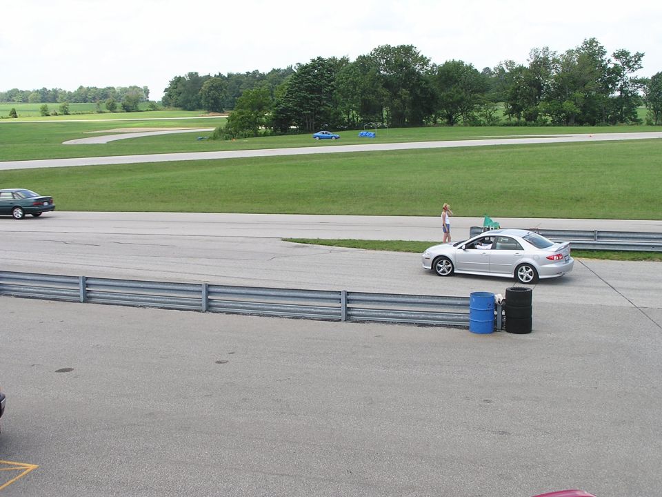 Silver sedan with a spoiler on a racetrack, a woman stands nearby. Another car is on the track in the distance