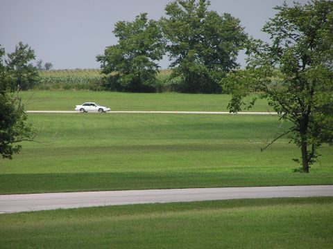 1993 Ford Taurus - A white sedan travels down a road cutting through a wide, green field under a hazy sky.