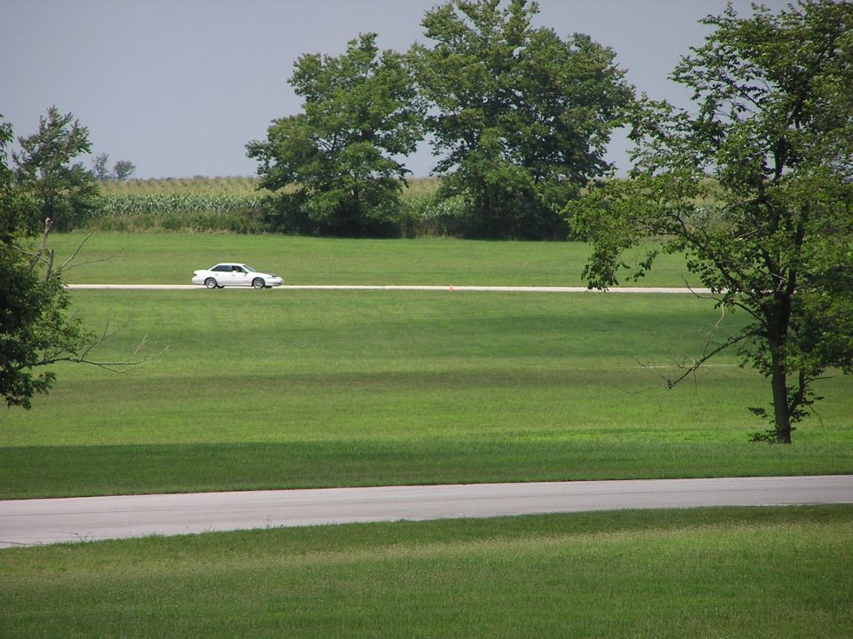 white sedan driving on a road through a grassy field with trees and corn in the background