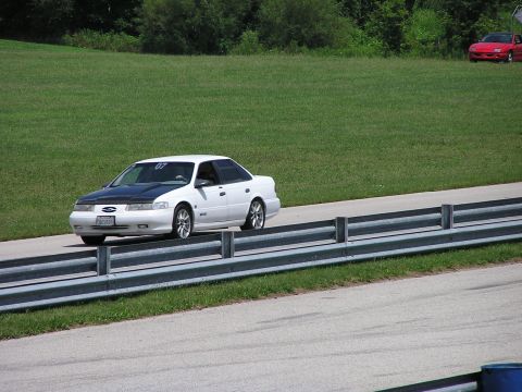 1993 Ford Taurus - A white 1993 Ford Taurus with a black hood and racing number 07 drives on a track, with another red car visible in the background.