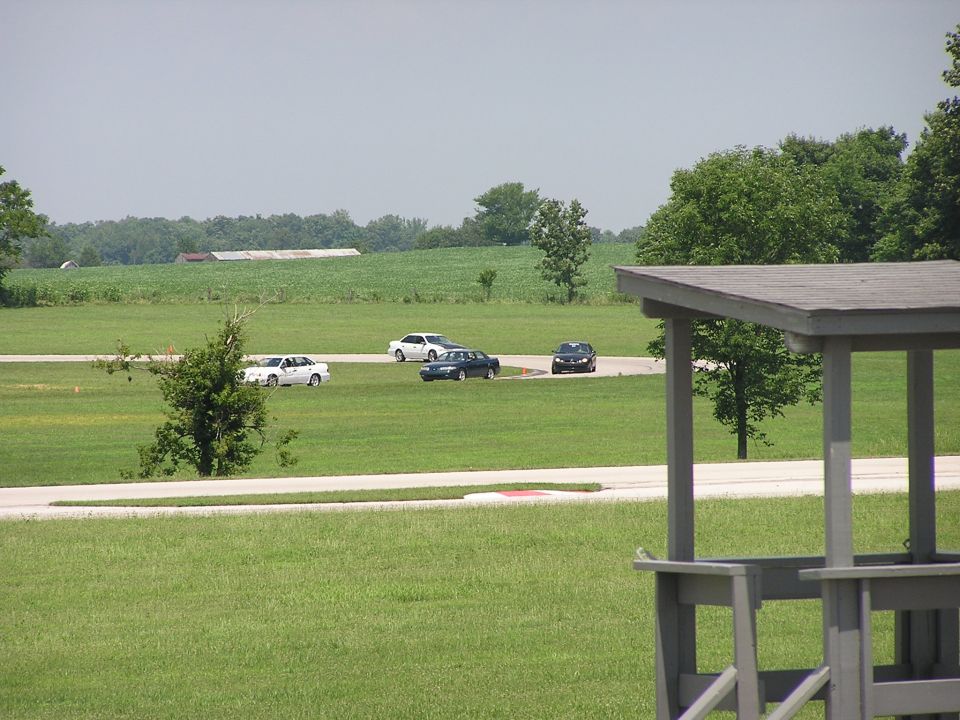 Several cars driving on a paved track in a rural area with fields and trees