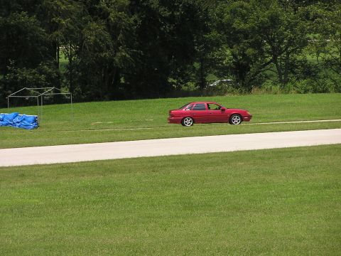 1993 Ford Taurus - A red 1993 Ford Taurus sedan is seen driving on a track with a grassy area and trees in the background.
