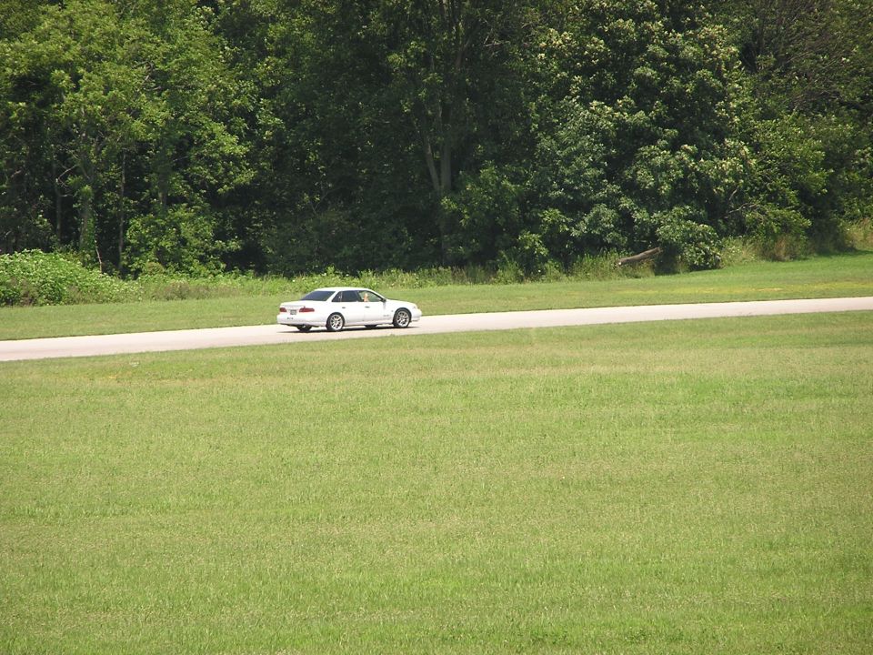 White sedan driving on a paved road with trees and grass in the background