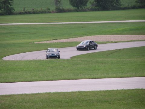 1993 Ford Taurus - Two sedans navigate a curve on a race track surrounded by green fields.