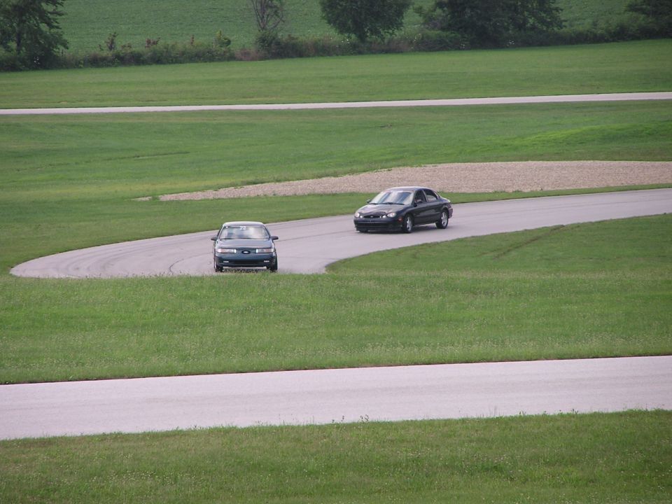 two sedans driving on a paved track with grassy areas around it
