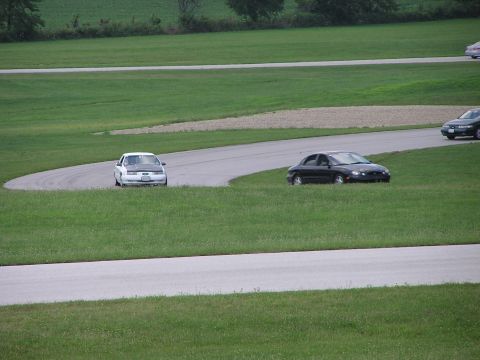1993 Ford Taurus - A white Ford Taurus with a black hood navigates a curve on a racetrack, with another car visible in the background.