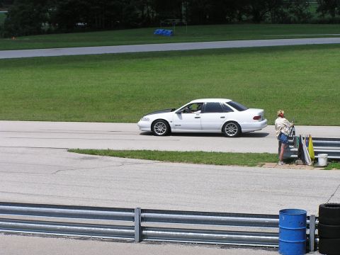 1993 Ford Taurus - A white 1993 Ford Taurus with a contrasting black hood is seen on a race track, with a person standing nearby holding a checkered flag.