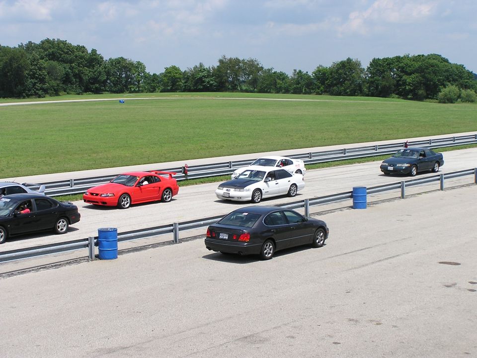 Dark gray sedan, possibly a Lexus GS, driving on a racetrack, rear 3/4 view
