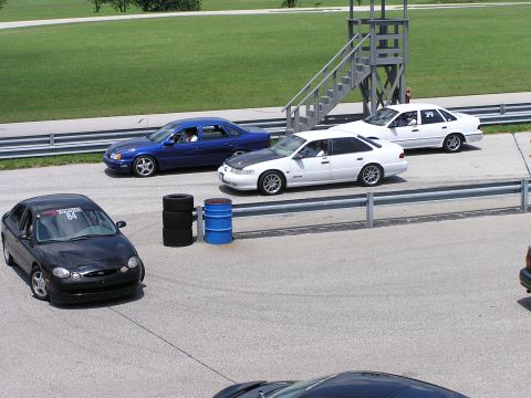 1993 Ford Taurus - A black 1993 Ford Taurus with racing numbers on its windshield is seen on a race track alongside other vehicles.