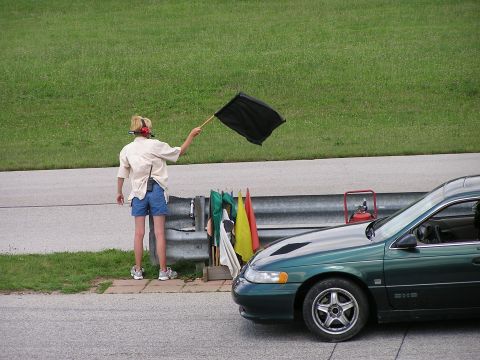 1993 Ford Taurus - A flag marshal holds a black flag next to a green sedan on a racetrack.