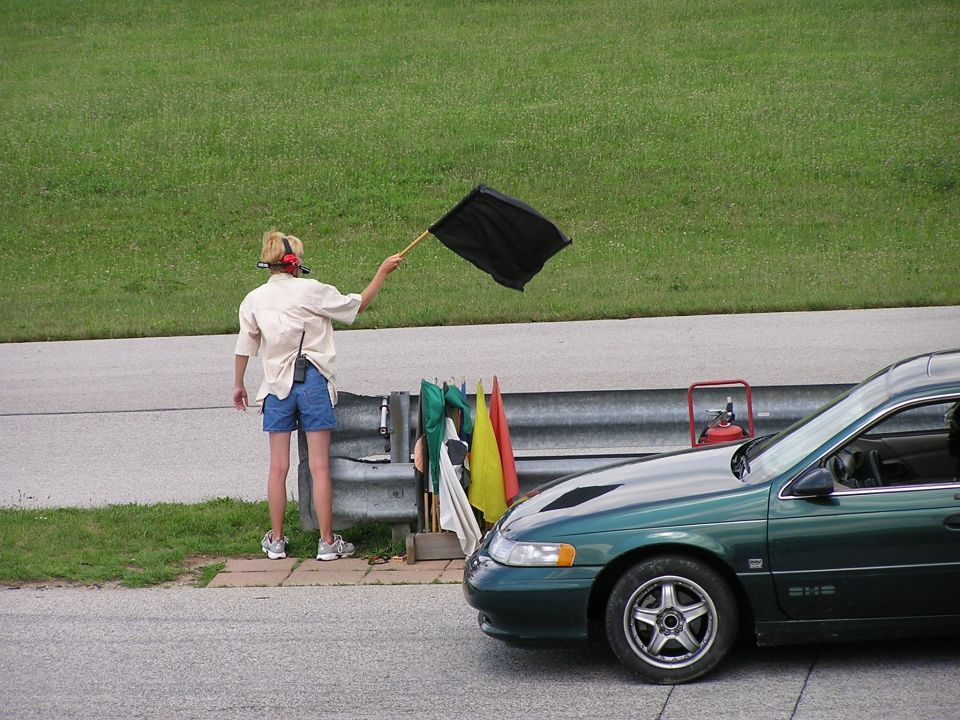 Green sedan parked on a racetrack with a flag marshal holding a black flag