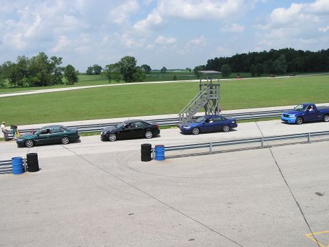 1993 Ford Taurus - A variety of vehicles are lined up on a racetrack, with a control tower in the background and trees beyond the grassy infield.