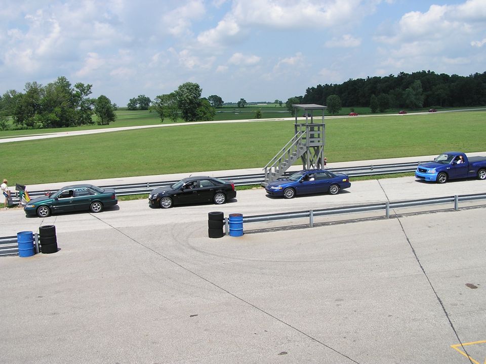 Four vehicles on a racetrack, including a green sedan, black sedan, blue sedan, and blue pickup truck