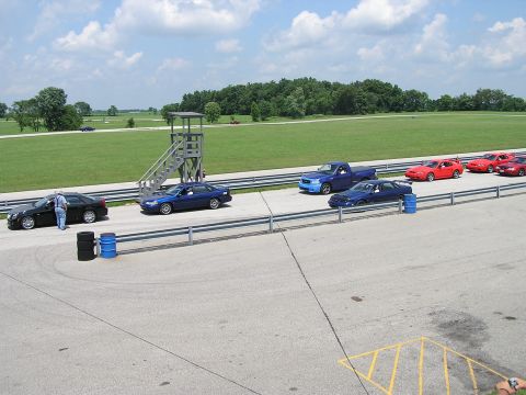 1993 Ford Taurus - Several vehicles are staged on a racetrack, with a tower and trees visible in the distance under a cloudy sky.
