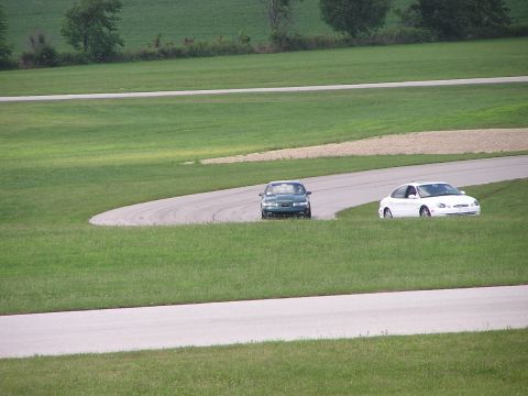 1993 Ford Taurus - Two sedans navigate a curve on a paved track surrounded by grassy hills.