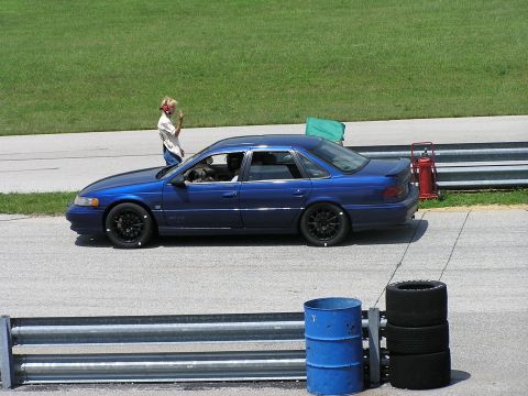 1993 Ford Taurus - A blue sedan is parked on the side of a racetrack with a person standing near the driver's side door.