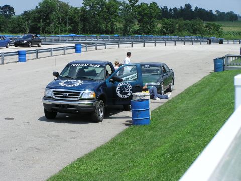 1993 Ford Taurus - A blue pickup truck and a green sedan are parked on the side of a race track, with people around them.