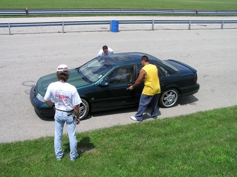 1993 Ford Taurus - Three men stand around a dark green sedan with a sunroof on a paved surface, possibly a racetrack.