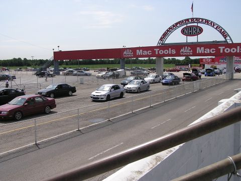 1993 Ford Taurus - Cars are parked and driving in the lot of a drag racing facility under a large archway.
