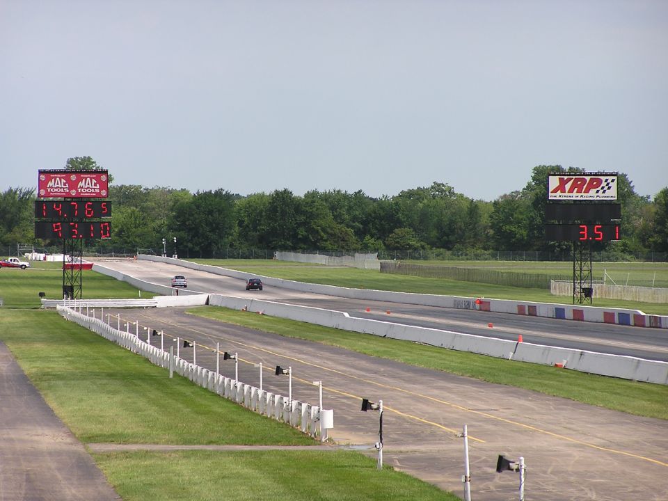Drag strip with two cars racing, scoreboards showing times. Trees and sky in background