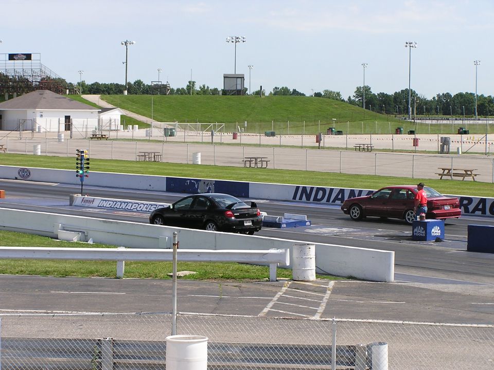 Two cars on a drag strip, one black and one red, with a starting tree and grandstands in the background