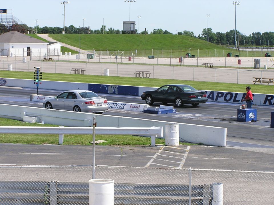 Two cars are lined up on a drag strip, one dark green sedan and one silver sedan. A person in a red shirt stands near the dark green car