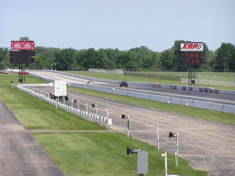 1993 Ford Taurus - Two cars compete on a drag strip under clear skies, with large digital scoreboards tracking their progress.