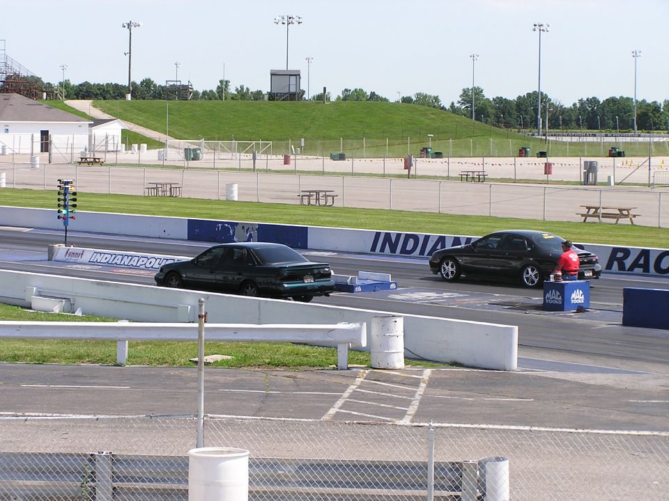 Two cars on a drag strip, one with a person in a red shirt standing nearby