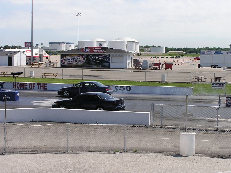 Two dark sedans race on a drag strip, with a building labeled 'Raceway Park' in the background