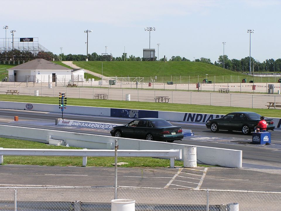 Two sedans on a drag strip at Indianapolis Raceway, with bleachers and buildings in the background