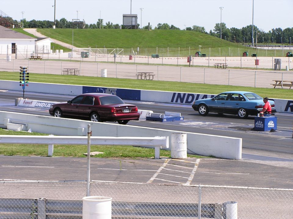 two cars at a drag strip, one red, one blue, with a person in red shirt next to the blue car