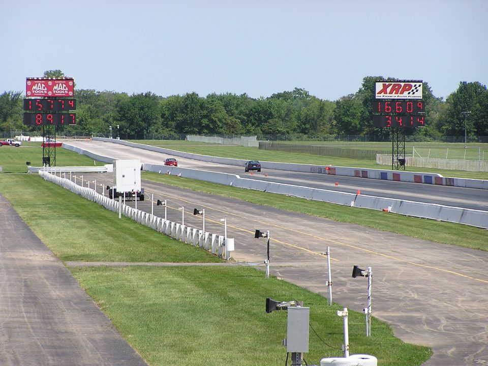Drag strip with two cars racing, scoreboards showing times, and trees in background