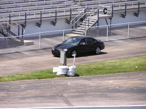 1993 Ford Taurus - A black sedan drives on a paved track with grassy areas and stadium seating visible.