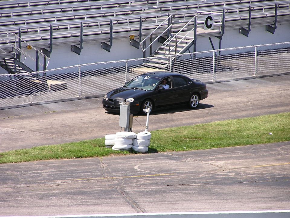 Black sedan driving on a track, with bleachers in the background. A number '107' is visible on the windshield