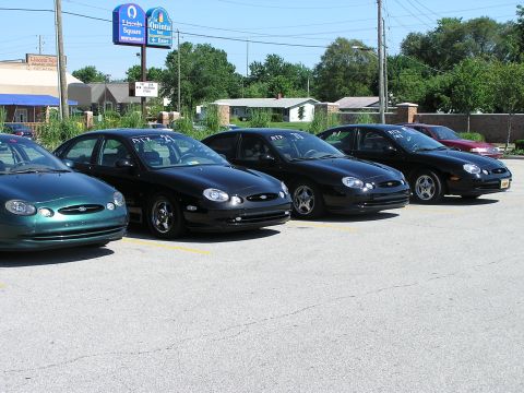 1993 Ford Taurus - Multiple Ford Taurus sedans are lined up in a parking lot under a bright sky.